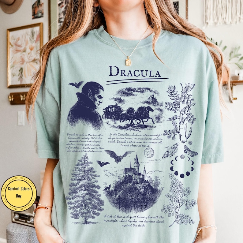 Op de afbeelding: Lichtgroen T-shirt met een donkerblauw grafisch ontwerp met het woord "DRACULA". Het ontwerp bevat illustraties van Dracula, een koets, een kasteel, vleermuizen en botanische elementen. Tekst is ook inbegrepen.