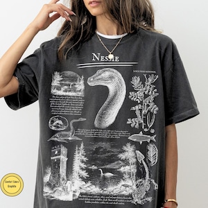 May include: A graphite gray t-shirt featuring a white graphic print of the Loch Ness Monster. The design includes the text "Nessie" and "Loch Ness Monster" alongside detailed illustrations of the creature, a stone tower, trees, and fish.