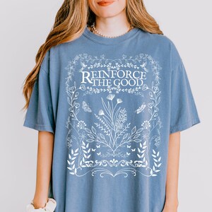 May include: A light blue t-shirt with a white floral design and the text "Reinforce the Good" in a white banner.
