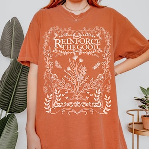 May include: A rust-colored t-shirt with a white floral design and the text "Reinforce the Good".