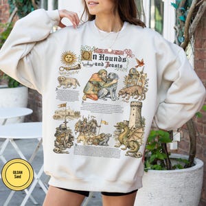 May include: Cream-colored sweatshirt with a medieval-themed graphic. The design includes illustrations of dogs, a dragon, a knight, and text that reads "Hounds and Beasts." The sweatshirt has a crew neck and long sleeves.