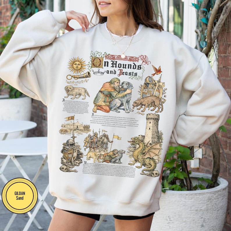 May include: Cream-colored sweatshirt with a medieval-themed graphic. The design includes illustrations of dogs, a dragon, a knight, and text that reads "Hounds and Beasts." The sweatshirt has a crew neck and long sleeves.