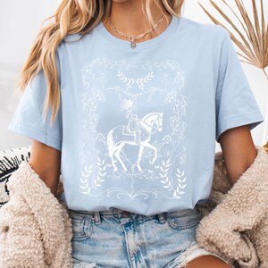 May include: A light blue t-shirt with a white graphic of a woman riding a horse in a floral frame.
