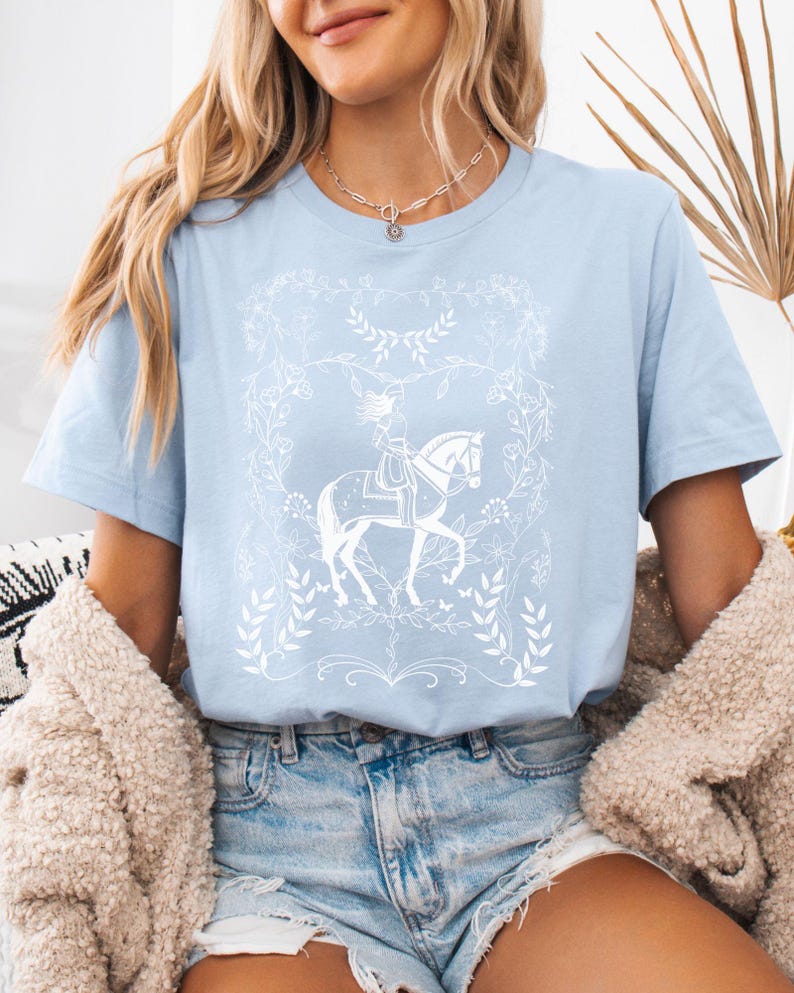 May include: A light blue t-shirt with a white graphic of a woman riding a horse in a floral frame.