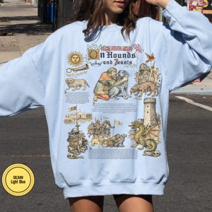 May include: Light blue sweatshirt with a medieval-themed graphic. The design includes illustrations of dogs, people, a dragon, and the text "In Hounds and Feasts". The sweatshirt has a crew neck and long sleeves.