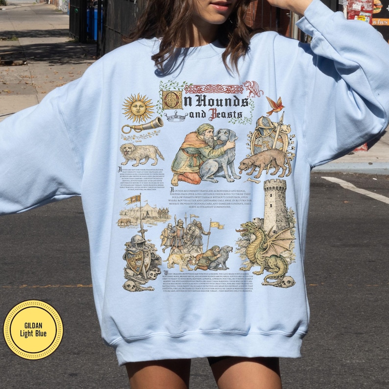 May include: Light blue sweatshirt with a medieval-themed graphic. The design includes illustrations of dogs, people, a dragon, and the text "In Hounds and Feasts". The sweatshirt has a crew neck and long sleeves.