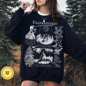 May include: Black sweatshirt with a white graphic design featuring the word "Frankenstein" at the top. The design includes an illustration of Frankenstein's monster, a wolf, a full moon, and botanical elements. The sweatshirt has a crew neck and long sleeves.