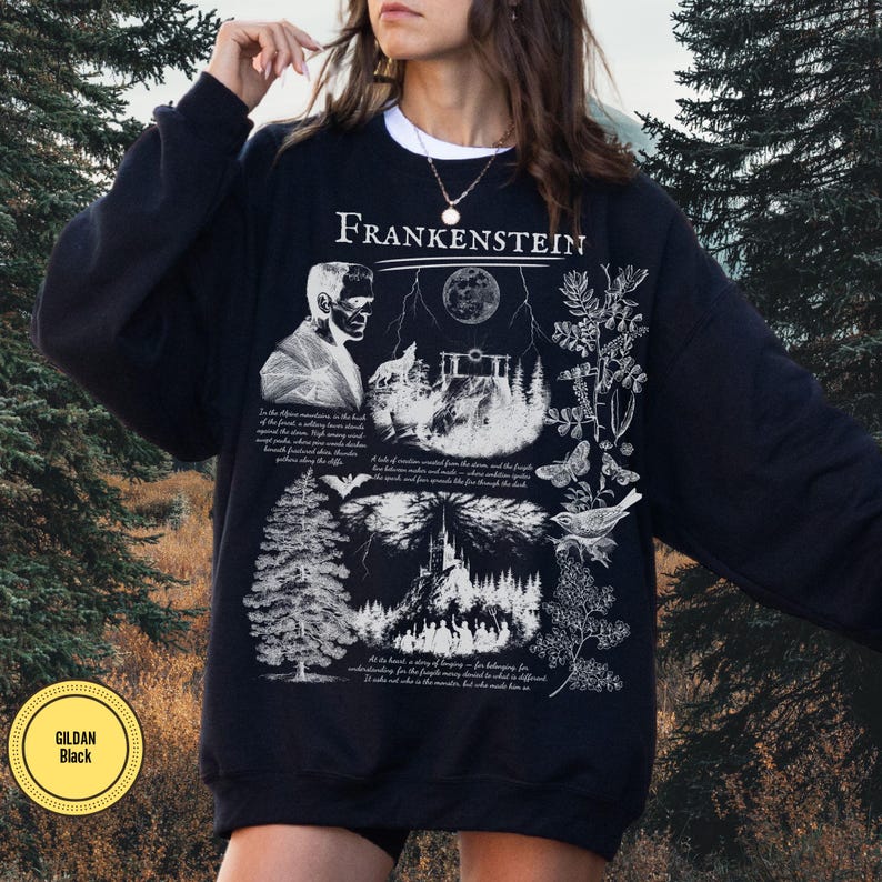 May include: Black sweatshirt with a white graphic design featuring the word "Frankenstein" at the top. The design includes an illustration of Frankenstein's monster, a wolf, a full moon, and botanical elements. The sweatshirt has a crew neck and long sleeves.
