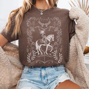 May include: A brown t-shirt with a white graphic of a woman riding a horse in a floral frame.
