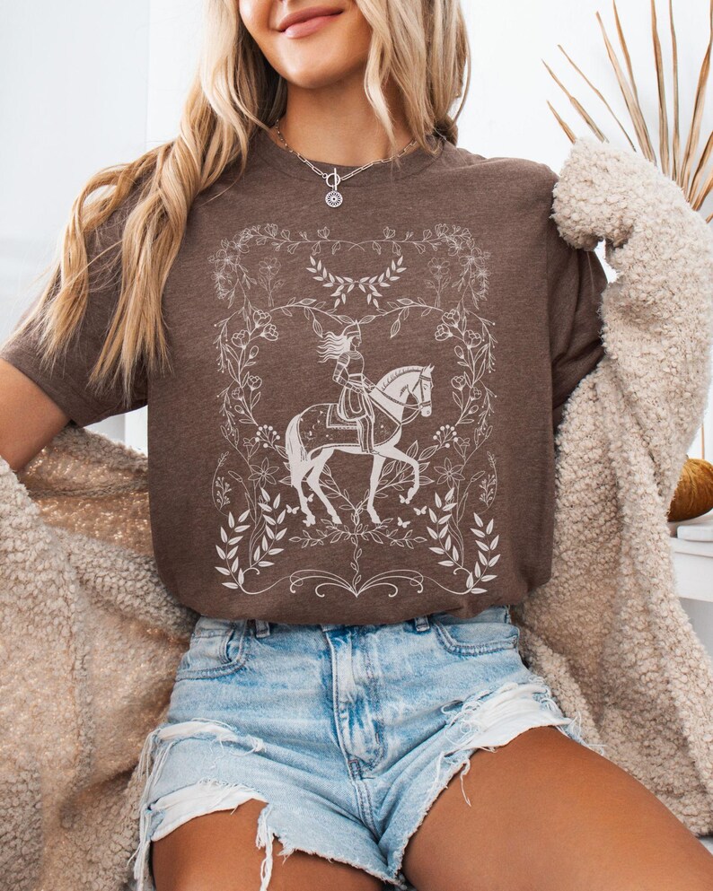 May include: A brown t-shirt with a white graphic of a woman riding a horse in a floral frame.