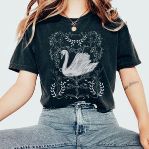 K&ouml;nnte beinhalten: Ein schwarzes T-Shirt mit einem wei&szlig;en Motiv eines Schwans in einem Blumenkranz. Der Schwan ist nach links gerichtet und hat einen langen Hals und einen gebogenen Schnabel. Der Blumenkranz besteht aus Bl&auml;ttern und Blumen.