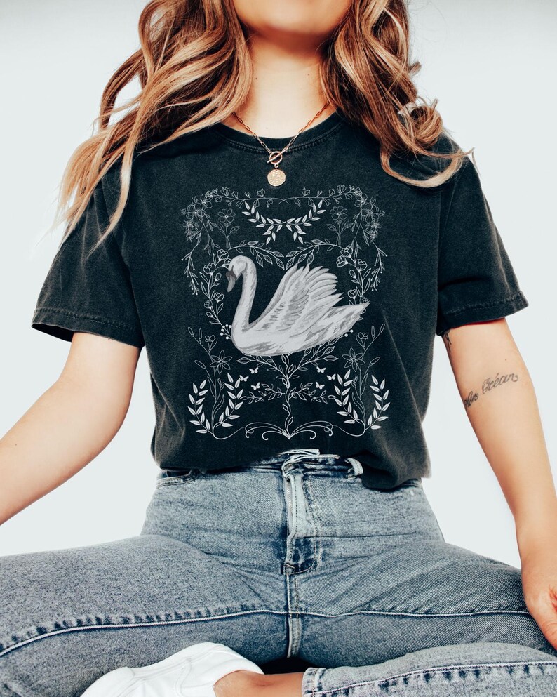 K&ouml;nnte beinhalten: Ein schwarzes T-Shirt mit einem wei&szlig;en Motiv eines Schwans in einem Blumenkranz. Der Schwan ist nach links gerichtet und hat einen langen Hals und einen gebogenen Schnabel. Der Blumenkranz besteht aus Bl&auml;ttern und Blumen.