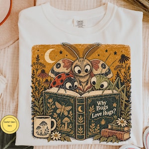 May include: An ivory t-shirt featuring a graphic print of a moth, a ladybug, and a grasshopper reading an open book titled Why Bugs Love Hugs? The illustration includes a crescent moon, stars, wildflowers, and a mug with mushroom designs.