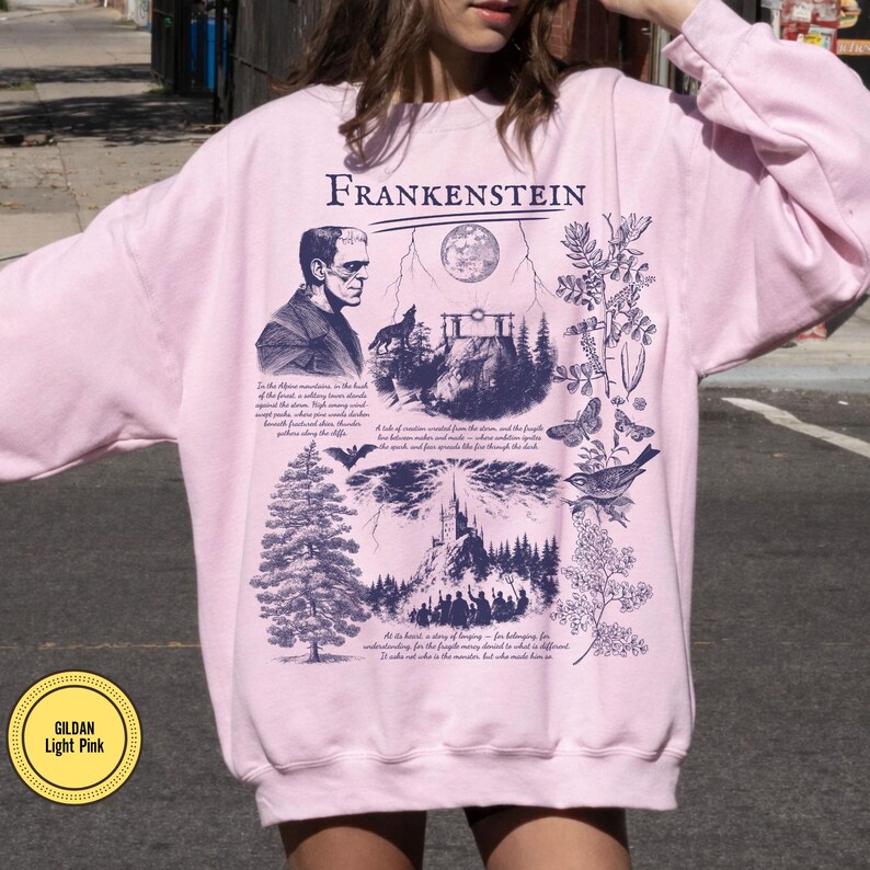 May include: Light pink sweatshirt with a graphic of Frankenstein, a portrait of the monster, and illustrations of a wolf, a full moon, a tree, a bat, a bird, and butterflies. The word "Frankenstein" is printed at the top.
