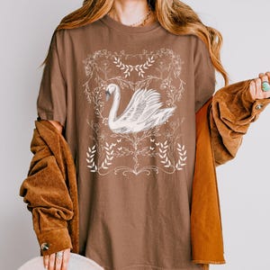 K&ouml;nnte beinhalten: Ein braunes Oversize-T-Shirt mit einer wei&szlig;en Grafik eines Schwans, umgeben von floralen Designs.