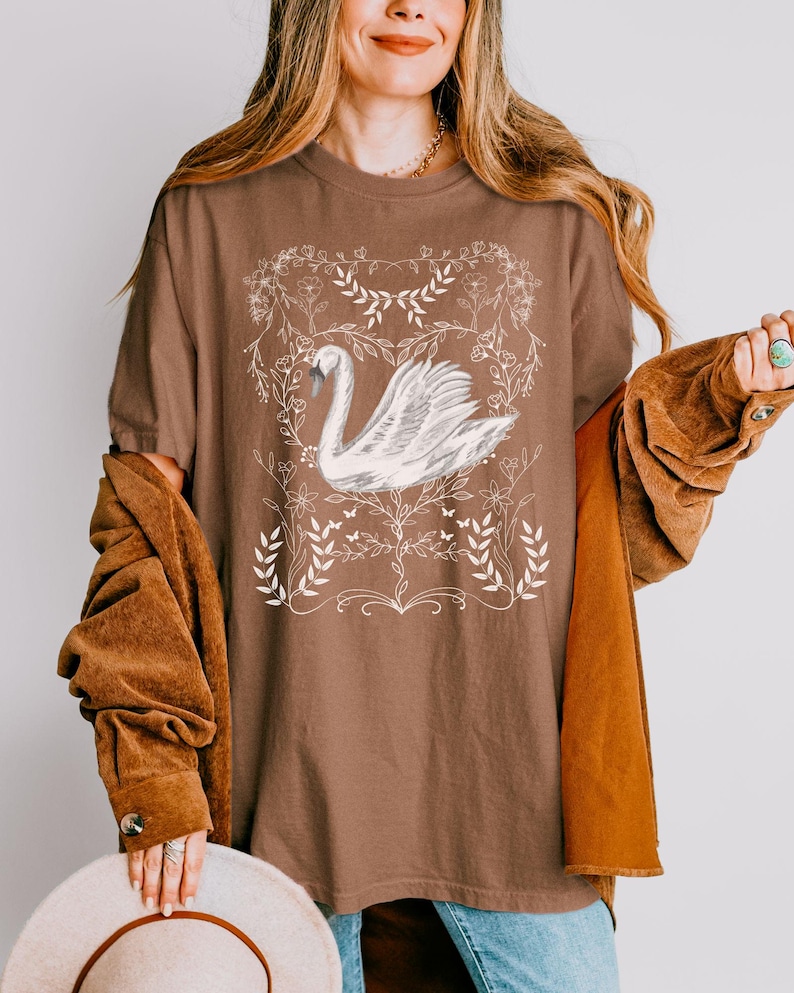 K&ouml;nnte beinhalten: Ein braunes Oversize-T-Shirt mit einer wei&szlig;en Grafik eines Schwans, umgeben von floralen Designs.