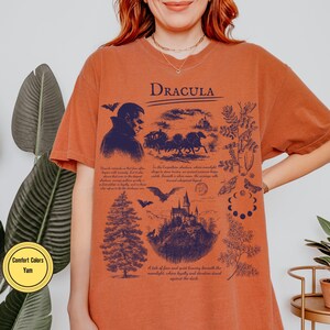 Op de afbeelding: Oranje Comfort Colors T-shirt met een donkerblauwe grafische print. Het ontwerp toont het woord "DRACULA", een vampier, een koets, een kasteel en tekst. Het T-shirt heeft een relaxte pasvorm.