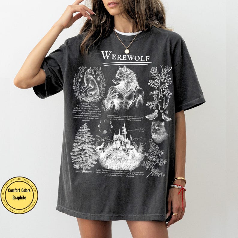 May include: A graphite-colored short-sleeve t-shirt featuring a white graphic print. The design includes the word Werewolf, illustrations of a wolf, a castle, a pine tree, a moth, and lunar phases, accompanied by small blocks of descriptive text.