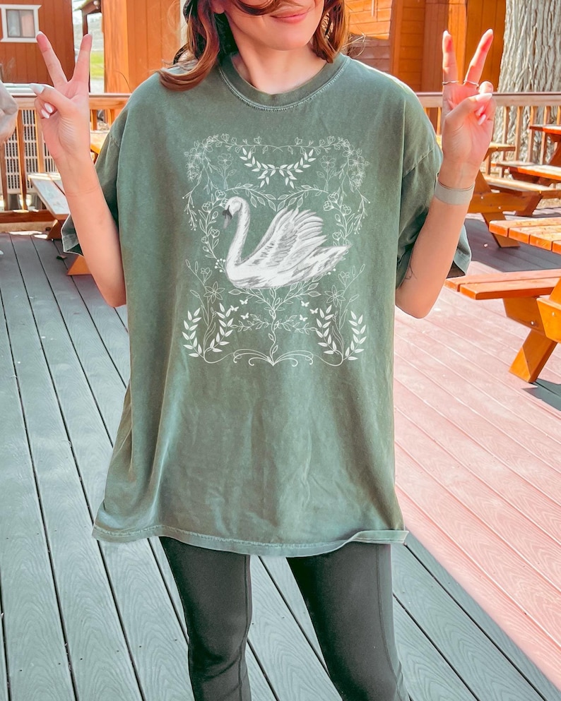 May include: A green oversized t-shirt with a white graphic of a swan in a floral wreath design.