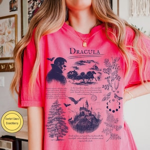 Op de afbeelding: Een roze t-shirt met het woord "Dracula" in het zwart gedrukt. Het shirt heeft een afbeelding van een vampier, een paardenkoets, een kasteel en botanische illustraties. De tekst "Comfort Colors Crunchberry" staat in een gele cirkel.