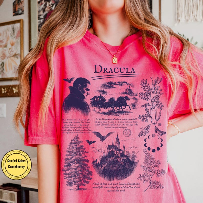 Op de afbeelding: Een roze t-shirt met het woord "Dracula" in het zwart gedrukt. Het shirt heeft een afbeelding van een vampier, een paardenkoets, een kasteel en botanische illustraties. De tekst "Comfort Colors Crunchberry" staat in een gele cirkel.