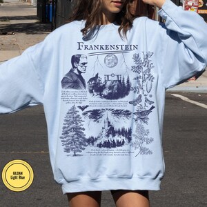 May include: Light blue sweatshirt featuring a graphic design with the word "Frankenstein" at the top. The design includes an illustration of Frankenstein's monster, a wolf, a full moon, a tree, and other gothic elements. The sweatshirt is a Gildan Light Blue.