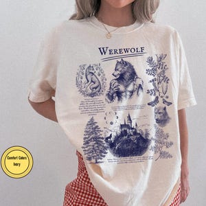 May include: An ivory t-shirt featuring a dark blue graphic print with the text "WEREWOLF" at the top. The design includes illustrations of a werewolf, a wolf, a castle, a pine tree, moon phases, and botanical elements with descriptive text.