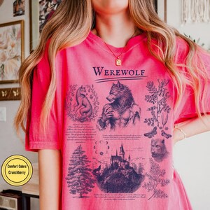 May include: A bright pink t-shirt featuring a dark blue graphic print with the text "WEREWOLF" at the top. The design includes illustrations of a werewolf, a wolf, a castle, a pine tree, moon phases, and botanical elements with small text.
