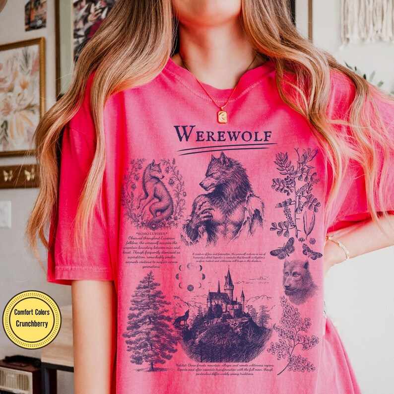 May include: A bright pink t-shirt featuring a dark blue graphic print with the text "WEREWOLF" at the top. The design includes illustrations of a werewolf, a wolf, a castle, a pine tree, moon phases, and botanical elements with small text.