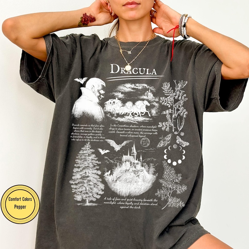 Op de afbeelding: Donkergrijs t-shirt met een grafisch ontwerp met het woord "Dracula". Het ontwerp bevat illustraties van een vampier, een paardenkoets, een kasteel, vleermuizen en botanische elementen. Het shirt is gelabeld "Comfort Colors Pepper".