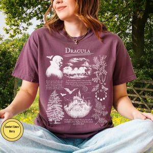 Op de afbeelding: Bessenkleurig T-shirt met een grafisch ontwerp met het woord "Dracula" bovenaan. Het ontwerp bevat illustraties van een vampier, een koets, een kasteel, vleermuizen en botanische elementen. Het shirt is van het merk Comfort Colors.