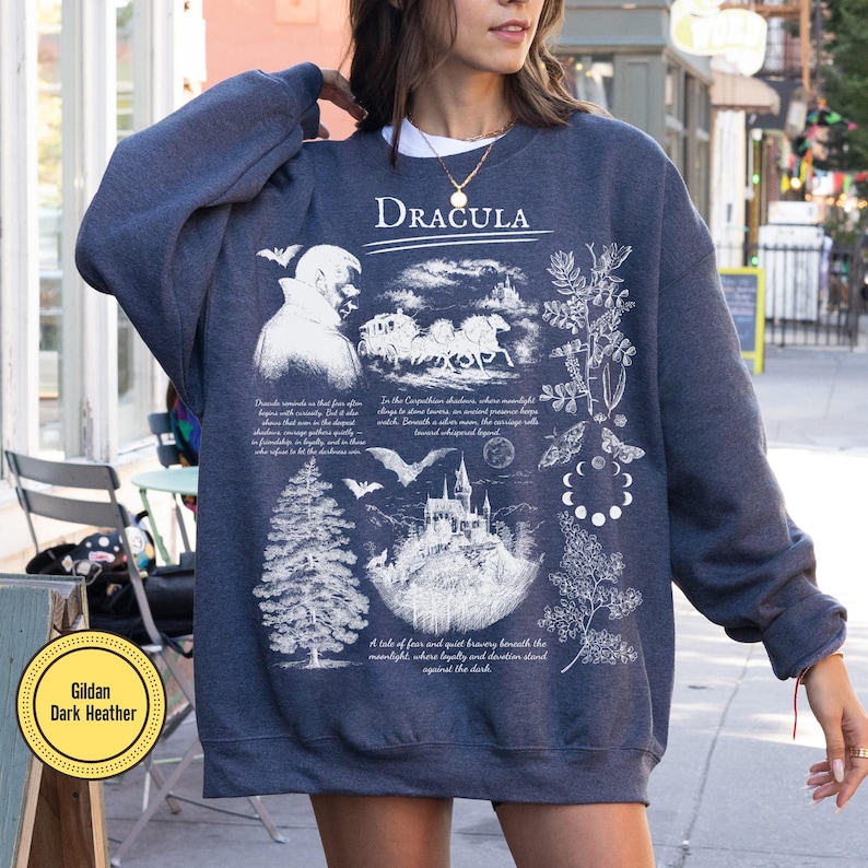 Op de afbeelding: Donkergrijze sweatshirt met een witte grafische opdruk met het woord "Dracula", een kasteel, vleermuizen en botanische illustraties. De sweatshirt is een Gildan Dark Heather.