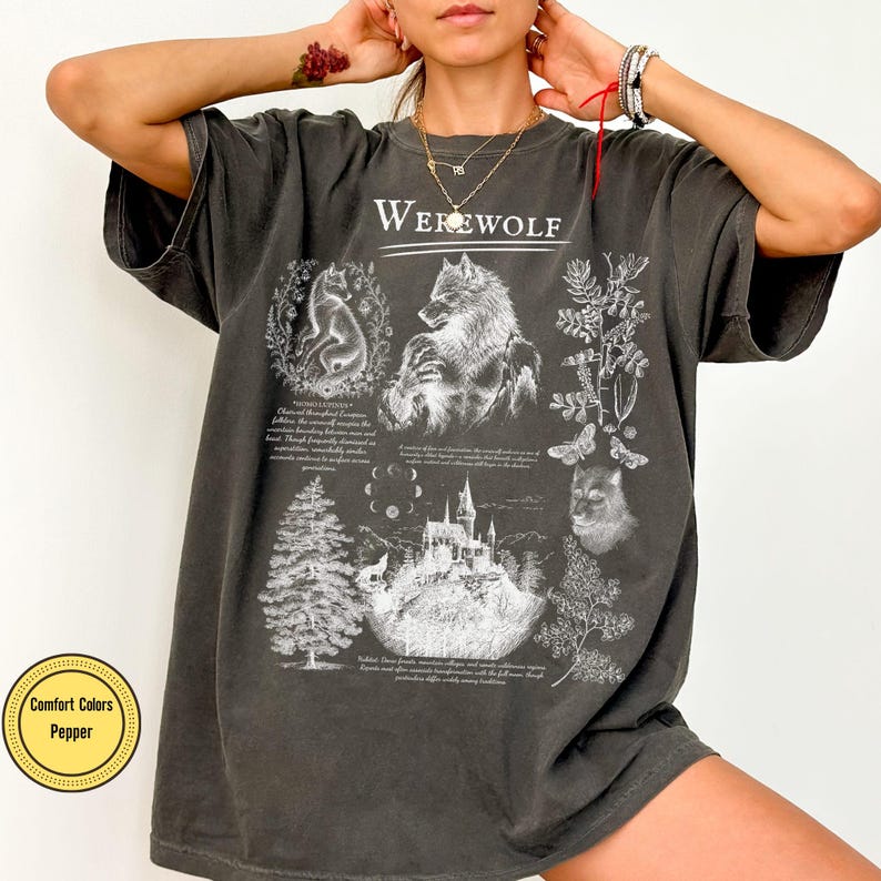 May include: A charcoal gray t-shirt featuring a white graphic print with the text "WEREWOLF" at the top. The design includes illustrations of a wolf, a castle, a pine tree, botanical elements, and moon phases, accompanied by small blocks of text.
