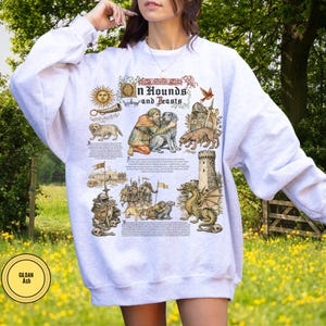 May include: A white sweatshirt featuring a medieval-themed graphic with the text "On Hounds and Beasts." The design includes illustrations of dogs, people, a dragon, and a castle. The Gildan Ash label is visible.