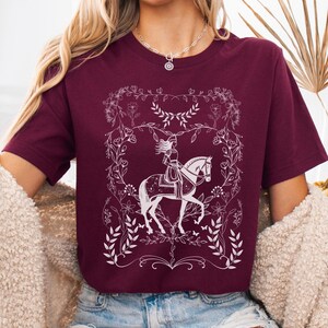 May include: A maroon t-shirt with a white graphic of a woman riding a horse in a floral frame.