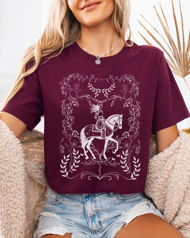 May include: A maroon t-shirt with a white graphic of a woman riding a horse in a floral frame.