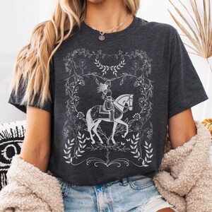 May include: A dark gray t-shirt with a white graphic of a woman riding a horse in a floral frame. The graphic is stylized and has a vintage feel.