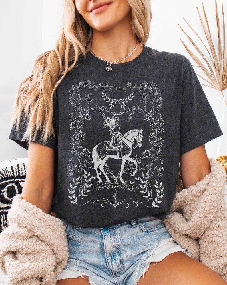 May include: A dark gray t-shirt with a white graphic of a woman riding a horse in a floral frame. The graphic is stylized and has a vintage feel.