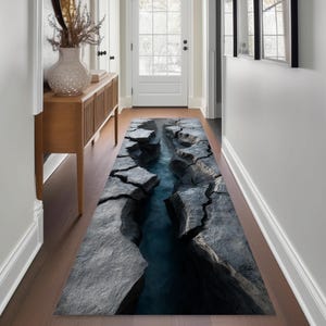 May include: A long, rectangular rug with a 3D image of a rocky canyon with a blue river running through it. The rug's design features gray rock formations on either side of the water, creating a realistic and textured appearance. The rug is placed in a hallway.