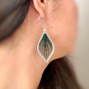 Può includere: Un orecchino argentato a forma di foglia. L'orecchino presenta un motivo a filo verde scuro all'interno di una cornice argentata. È sospeso a un gancio argentato. Il design è visibile su uno sfondo sfocato.