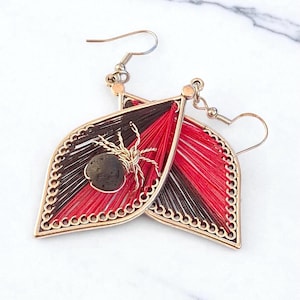 May include: A pair of handmade leaf-shaped earrings. The earrings feature a gold-colored frame with a decorative edge. The interior is filled with red, black, and brown thread, and a small spider and stone are attached.