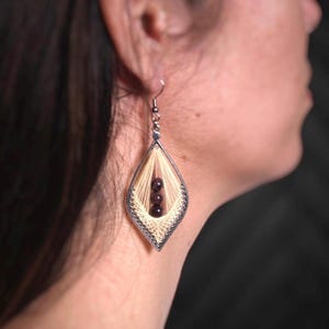 May include: A silver-toned, teardrop-shaped earring with a cream-colored woven design and three dark red beads. The earring is worn on a person's ear, with the focus on the intricate details of the jewelry.