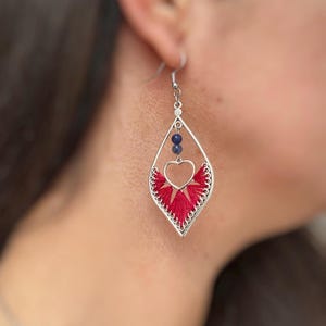 May include: Silver-toned dangle earrings with a red embroidered fan design and a heart-shaped cutout. The earrings feature two small blue beads and a decorative edge. A stylish accessory.