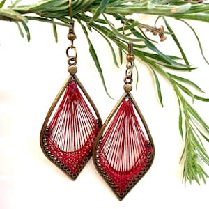 Può includere: Un paio di orecchini in filo rosso con una cornice in metallo color bronzo. Gli orecchini hanno la forma di una lacrima con un design delicato e intricato.