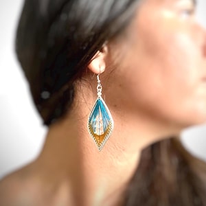 以下が含まれることがあります： 青と金のデザインが施された、銀色のしずく型のイヤリング。イヤリングは、銀色のフレームの中に繊細な織り模様が施されています。イヤリングはシンプルなフックから吊り下げられています。