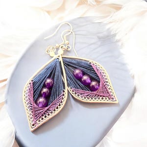 May include: A pair of leaf-shaped earrings with a silver-tone frame. The earrings feature navy blue and purple threadwork, with three purple beads in each leaf. The edges are trimmed with a decorative purple thread.