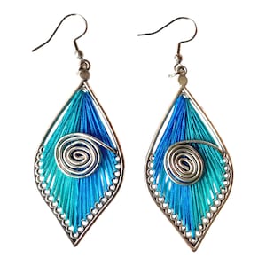 May include: A pair of handmade earrings with a teardrop shape. The earrings feature a silver-colored metal frame filled with blue and teal thread. A silver spiral design is centered on each earring, and the edges are lined with small silver beads.