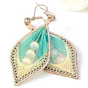 May include: A pair of teardrop-shaped earrings featuring a gold-toned metal frame with intricate cutouts. The center is filled with a geometric web of teal and cream-colored thread, accented by three smooth, round, translucent white beads.