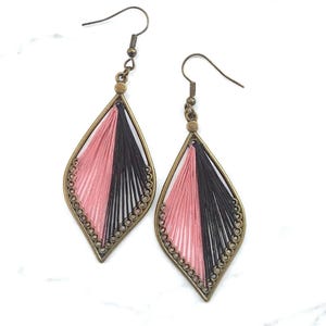May include: A pair of handmade earrings with a geometric design. Each earring features a teardrop-shaped frame in a bronze tone, filled with pink and black thread. The earrings have a beaded border and a hook closure.
