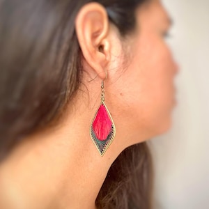 Può includere: Un singolo orecchino pendente a forma di goccia. L'orecchino presenta un vivace centro ricamato rosa, incorniciato da una filigrana in metallo color oro. La parte superiore dell'orecchino è di colore verde scuro. L'orecchino è appeso a un gancio color oro.